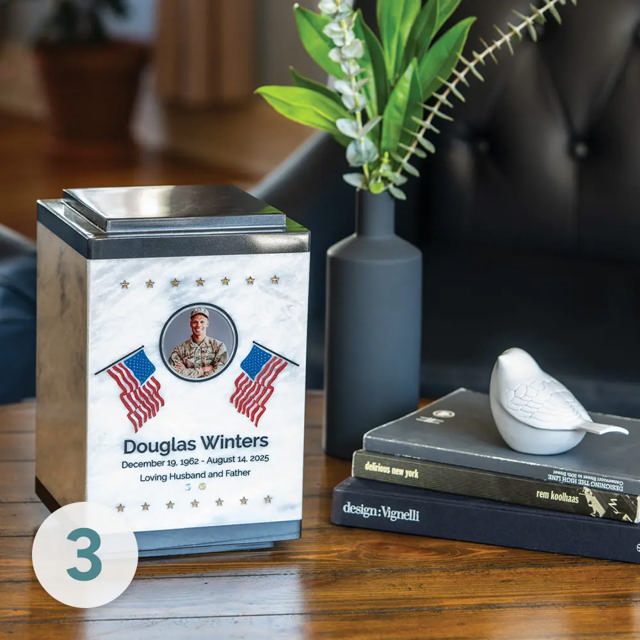 A white marble urn with an American flag design sits on a table next to books and a bird keepsake